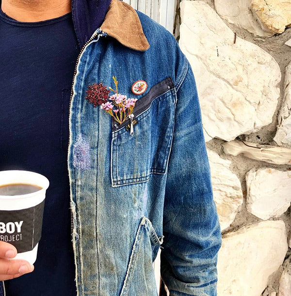 Closeup of torso in jean jacket with navy undershirt. Flower in breast pocket. Holding cup of Flowerboy Coffee